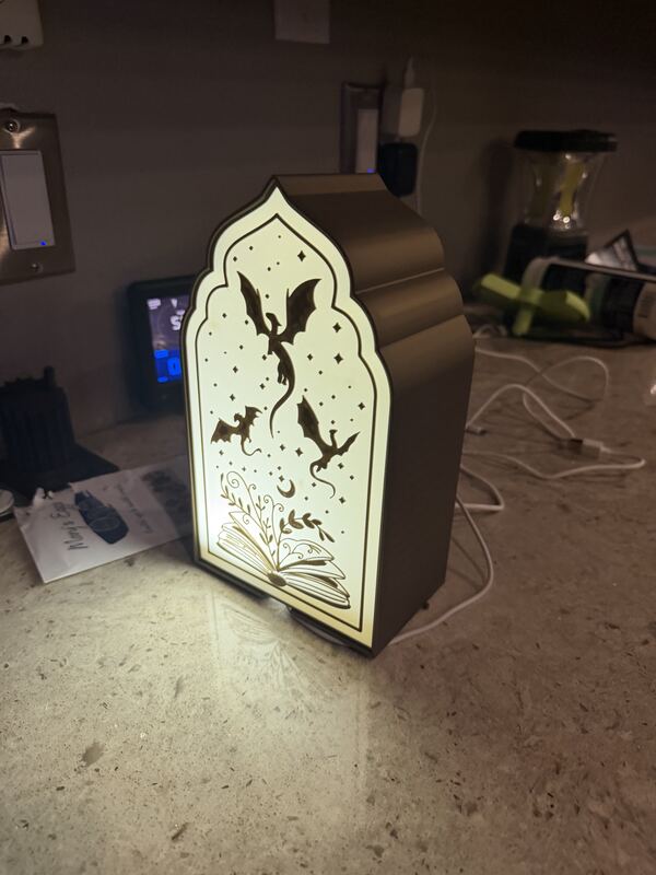 Illuminated 3D printed lamp with a bat and leaf pattern cut into the shade, glowing on a bookshelf.