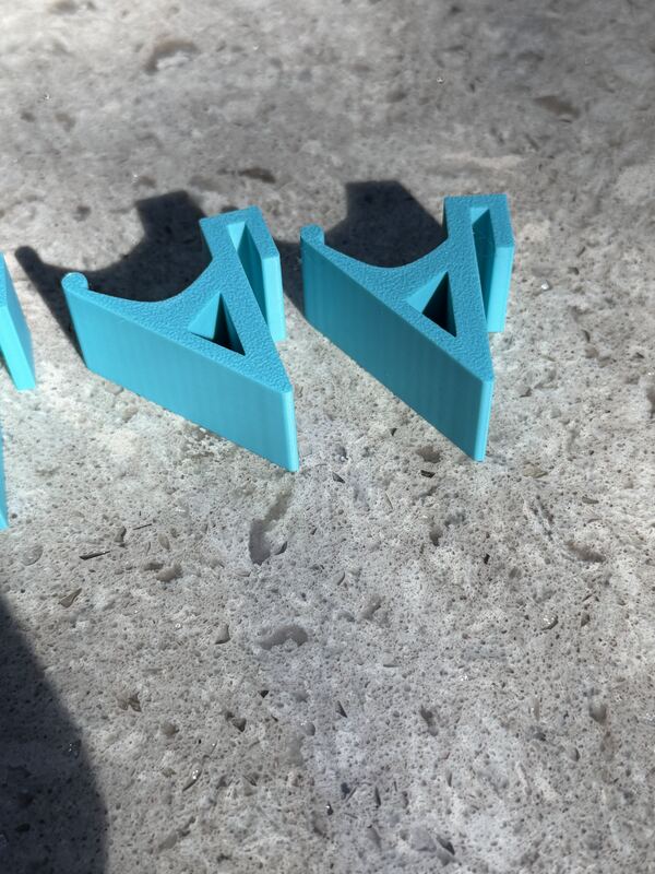 Two cyan 3D printed angle brackets with triangular gusset cut-outs resting on concrete.